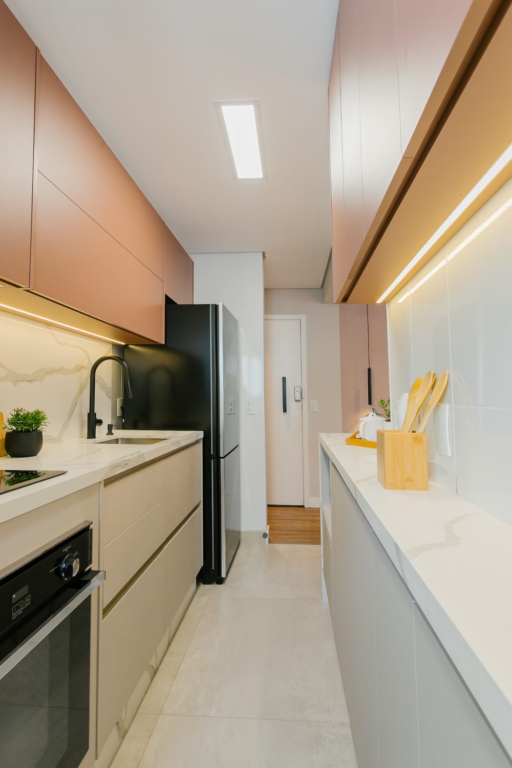 Projeto RBC: cozinha corredor com armários superiores rosé iluminados por LED, bancadas brancas com veios e geladeira ao fundo